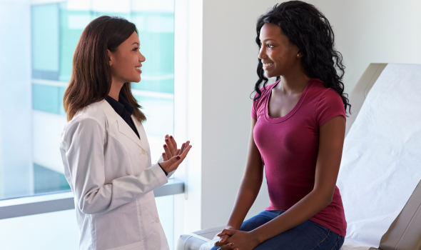 Female doctor in a white coat chats with a smiling patient seated on an exam table in a bright clinic.