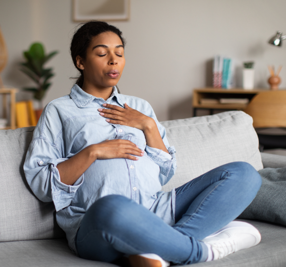 Pregnant woman sitting on a sofa with eyes closed, one hand on her chest and the other on her baby bump.