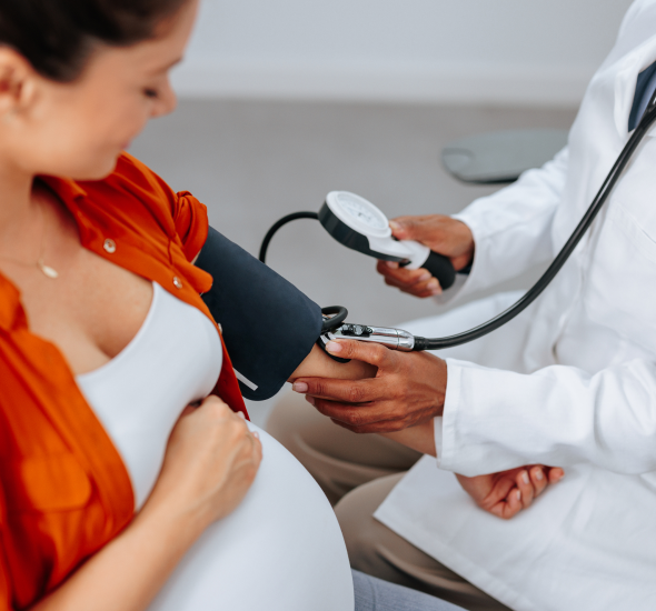 Pregnant woman in orange shirt has her blood pressure checked by a clinician using a cuff and stethoscope on her arm.