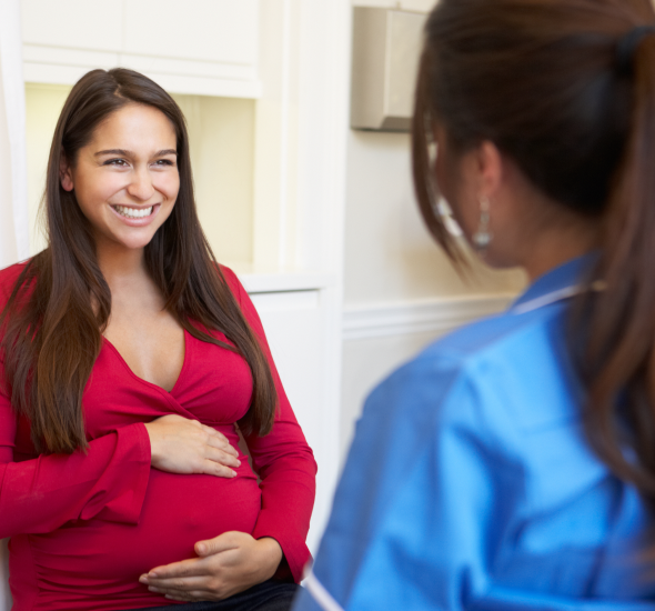 Pregnant woman in a red top smiles during a prenatal check-up with a clinician in blue scrubs in a bright clinic.