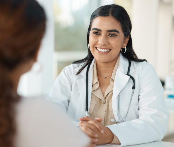 Female doctor in a white coat smiles while talking with a patient at a clinic, hands folded on the table, stethoscope around her neck.